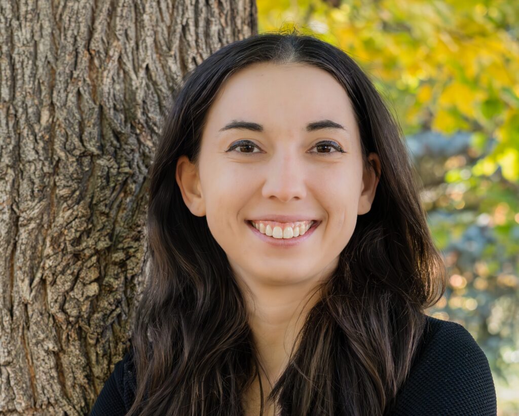 A photo of Erin J.K. Truesdell, a white woman in her 20s with brown eyes, brown hair, and winged eyeliner. She is smiling.
