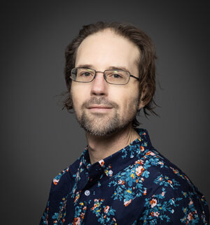 Headshot of Travis Faas, an individual with light skin, brown hair, and glasses, on a solid background.